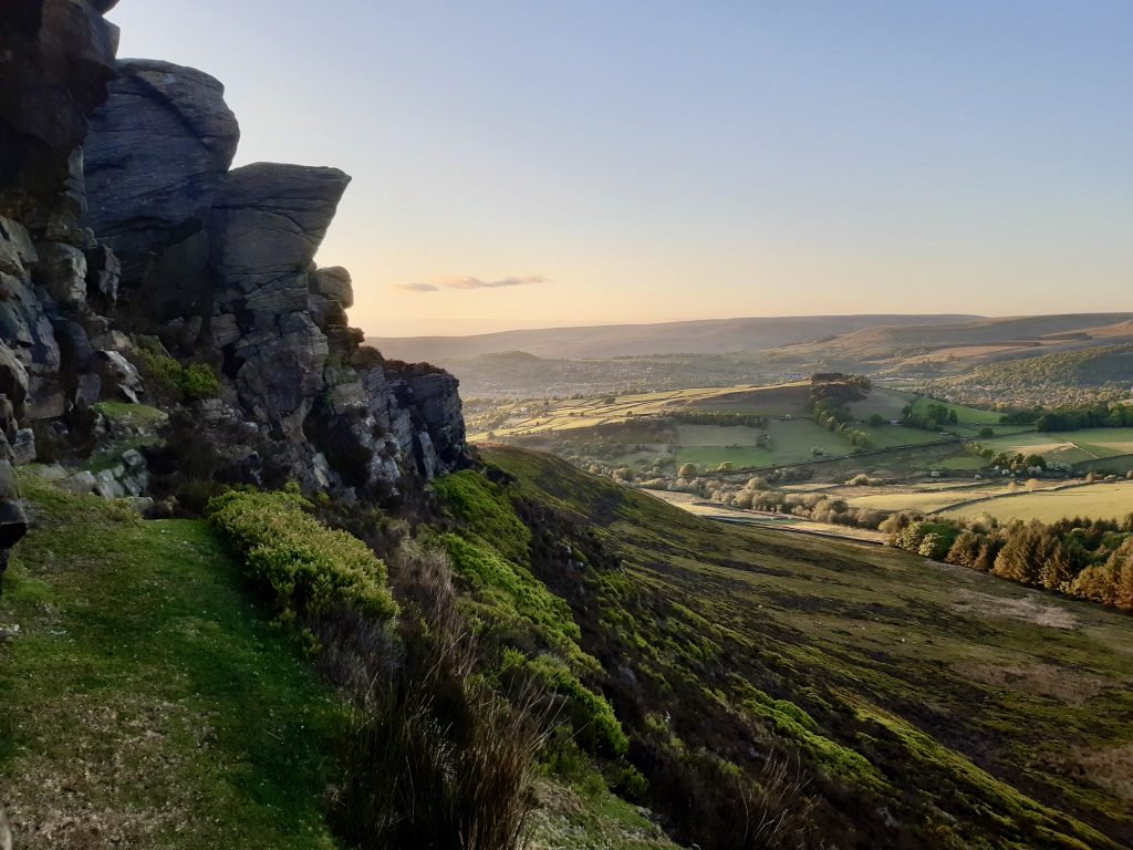 Hill day in the Peak District with a gritstone edge and a hillside gently sloping down in the foreground with softly lit fields in the distance.